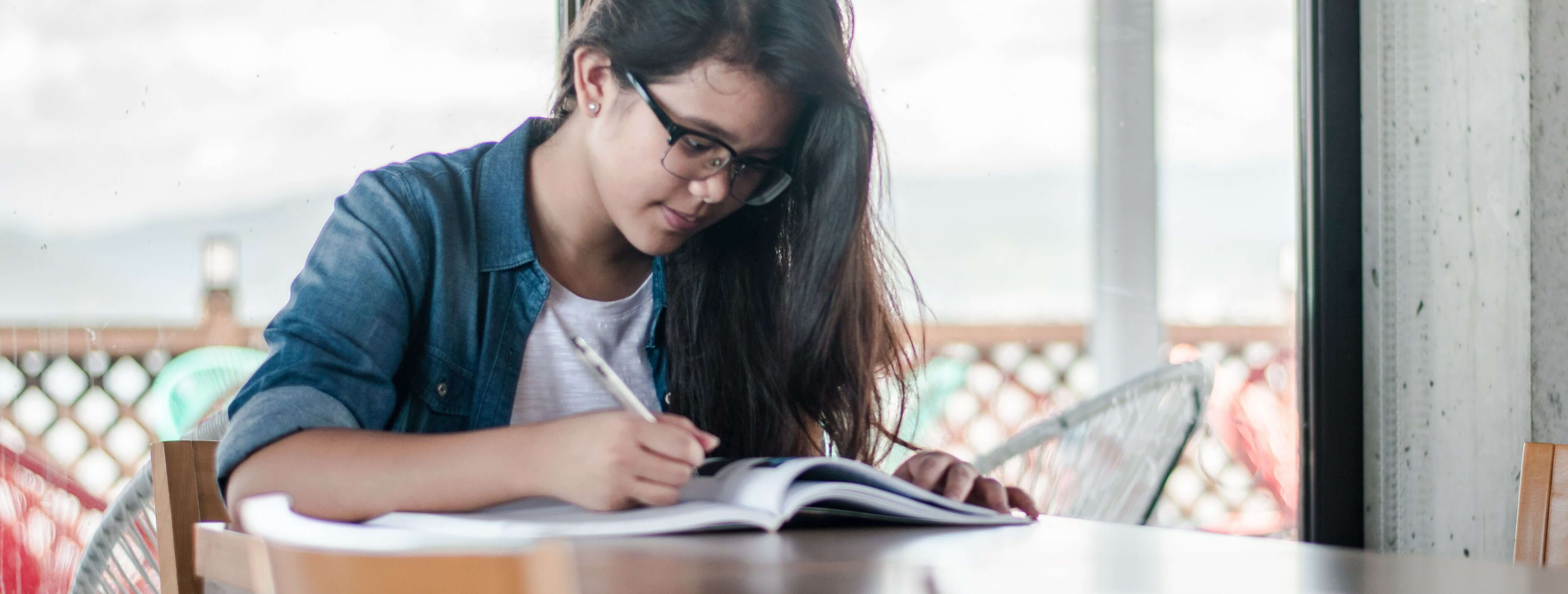 Imagem de uma menina olhando para um livro aberto na mesa e segurando um lápis.
